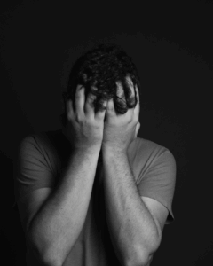black-white photo: sad man with curly hair puts his hands in front of his face to hide his agony.